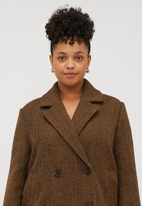 Cappotto doppiopetto in tessuto a scacchi marrone e nero con grandi rever, texture liscia e tasche laterali. Bottoni neri sono visibili.
