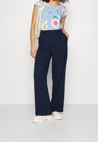 Top floreale blu con maniche corte, abbinato a pantaloni a gamba larga blu navy. Modella che indossa scarpe platform bianche e porta una borsa leggera.