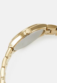 Reloj tono dorado con una caja suave y redondeada y pulsera enlazada; cuenta con un botón del logo de CK y un acabado pulido en las superficies metálicas.