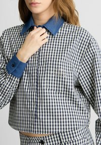 Karierter Hemd mit einem navyblauen und weißen Gingham-Muster, Denim-Kragen und -Manschetten, einer Brusttasche und einem dekorativen Emblem auf der Tasche.