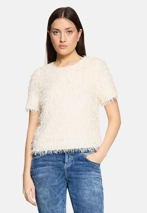 Fuzzy, cream-colored short-sleeve top with fringe detailing, paired with denim jeans. Casual cut; textured surface adds visual interest.