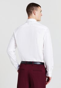 Camicia bianca a maniche lunghe con una texture liscia, abbinata a pantaloni bordeaux e una cintura nera, che mette in mostra un design pulito e aderente.