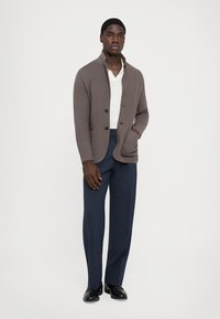 Gray cardigan with three buttons, textured fabric, and side pockets, worn over a light shirt and paired with dark trousers and shoes.
