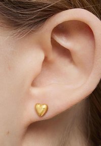 Orecchino a forma di cuore in oro, con superficie liscia, aderente al lobo dell'orecchio, che enfatizza un design lucido e uno stile minimalista.