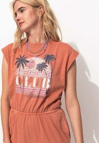 Combinaison sans manches couleur rouille avec taille élastique, ornée d'un imprimé graphique de palmiers et du texte "CLUB D'ÉTÉ" dans des couleurs claires et foncées.