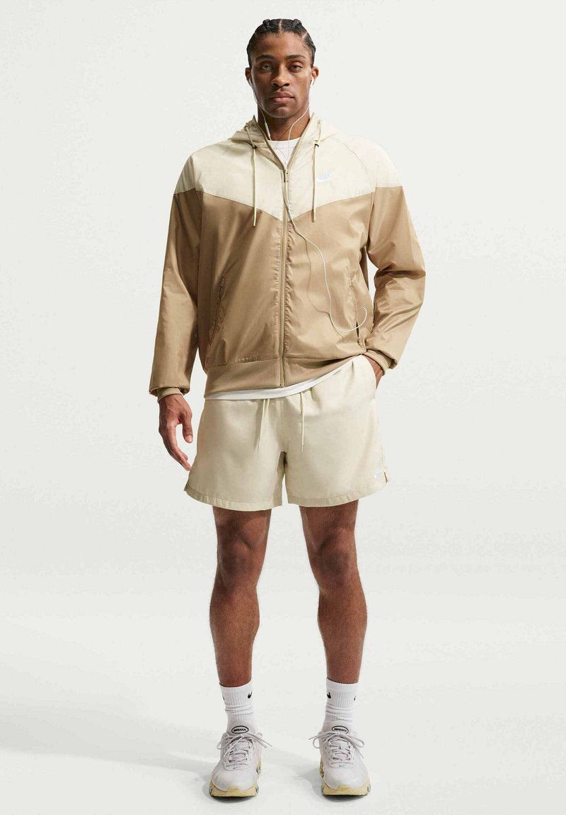 Man draagt een beige en crèmekleurige jas met rits, bijpassende korte broek, witte sokken, sneakers en bedrade oordopjes, staand tegen een effen achtergrond.