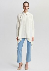 Chemise couleur crème avec un ourlet asymétrique, fermeture à boutons et coupe oversize. Associée à un pantalon en jeans bleu clair et des talons beiges.