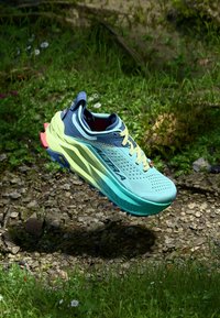 Pantofii sport aqua și bleumarin cu un superior din plasă, șireturi neon galbene și talpă cu tracțiune mare, suspendați deasupra unui fundal de iarbă și pietre.