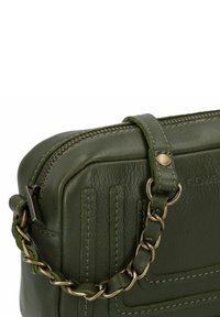 Primo piano di un angolo di borsa in pelle verde oliva con cerniera, dettagli cuciti e una tracolla a catena intrecciata con strisce di pelle.