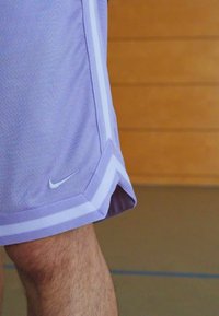 Lilla atletiske shorts lavet af åndbart mesh-stof, med et sort Nike-logo og hvide side-striber. Designet inkluderer en mønstret kant.