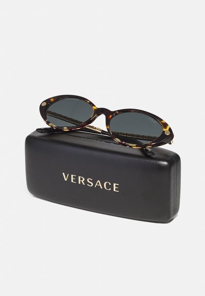 Kempinės akinių rėmeliai su ovaliais lęšiais, gulintys ant juodos dėžutės. Dėžutė turi auksinį užrašą "VERSACE". Sklandus paviršius, kompaktiškas dizainas.