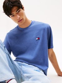 T-shirt in cotone blu con scollatura a girocollo che presenta una piccola patch con logo rosso, bianco e blu. Indossato con jeans chiari strappati.