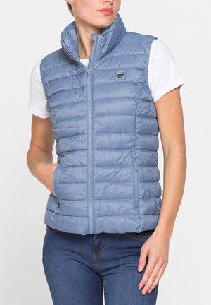 Donna che indossa un gilet trapuntato azzurro chiaro senza maniche sopra una maglietta bianca a maniche corte e jeans blu, con la mano nella tasca del gilet.
