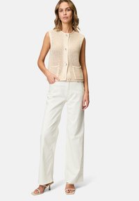 Beige strikvest med ærmeløs design, rund halsudskæring og forlommer. Sammen med hvide bukser med bred benvidde og lysebeige sandaler.