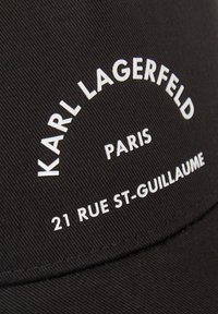 Svart keps av tyg med vit broderad text: "KARL LAGERFELD PARIS 21 RUE ST-GUILLAUME" i en bågdesign. Texturerad yta.