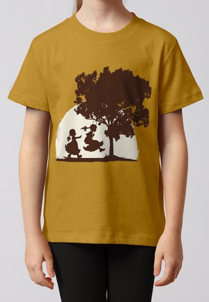 Kind trägt senfgelbes T-Shirt mit brauner Silhouette von zwei Mädchen, die auf einer Baum Schaukel spielen, vor cremefarbenem Kreis Hintergrund.