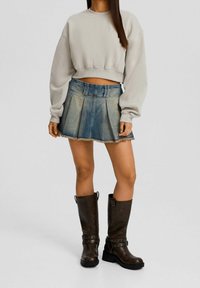 Kvinna som bär en kort beige sweatshirt, en plisserad denimminikjol med fransar och knähöga, mörkbruna läderstövlar med remmar.