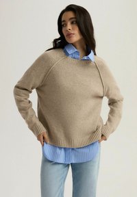 Beżowy sweter z dzianiny z prążkowanym wykończeniem i mankietami, nałożony na jasno-niebieską koszulę w paski. Proste tło, luźny fason, miękka tekstura.