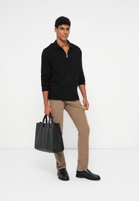 Maglione nero a maglia zip, pantaloni beige texture, scarpe nere slip-on e una borsa a tracolla in pelle nera con più scomparti.