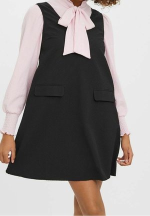 Femme portant une robe noire sans manches avec des poches avant par-dessus un chemisier rose clair à manches longues avec un grand col à nœud papillon.