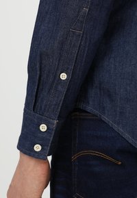 Tmavě denimová košile s dlouhými rukávy, s manžetou na knoflíky a jemnou texturou, s světle zbarvenými knoflíky a klasickým střihem.