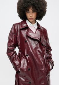 Trench-coat imperméable bordeaux à texture brillante, à design croisé, grand col et boutons noirs, porté par-dessus un pull léger.