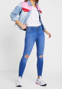 Jeansjacke mit pinken und blauen Farbblocks, weißem T-Shirt, roten blauen Jeans und weißen Sneakers mit pinken und blauen Akzenten.
