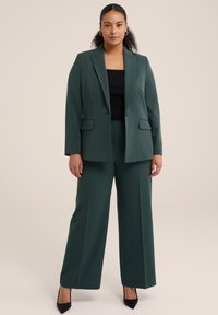 Tmavě zelený přizpůsobený blazer s klopami, dvěma předními kapsami, v kombinaci s širokými kalhotami ve stejném odstínu a černým topem. Nositelka v lodičkách.