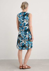 Ärmlös blå klänning med geometriska mönster i vitt, teal och brunt, med en lager-på-lager kjol och rund halsringning. Sandaler i brunt läder.