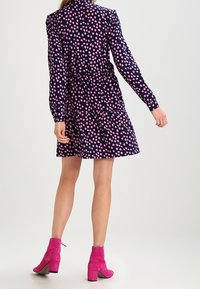 Navy-blaues gepunktetes Kleid mit rosa Mustern, langen Ärmeln, tailliertem Schnitt und ausgestelltem Rock. Kombiniert mit leuchtend rosa Wildleder-Stiefeletten.