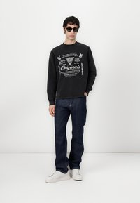 Sort sweatshirt med hvid grafisk print, blå jeans og hvide sneakers. Modellen har solbriller på og står foran en ensfarvet baggrund.