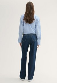 Camisa de botones a rayas azul y blanca con mangas largas, combinada con jeans de pierna ancha azul oscuro y zapatillas blancas, vista desde atrás.