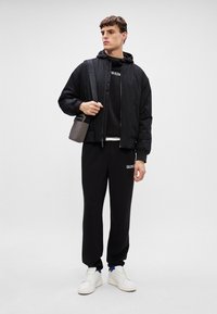 Czarna kurtka bomberka na czarnej bluzie z kapturem i dopasowanych dresach, z białymi sneakersami i małą szaro-skalną torbą na ramię. Minimalistyczny design.