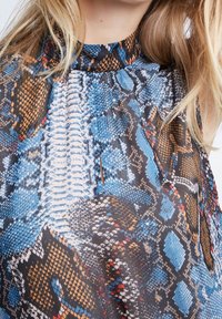 Blusa a cascata con colletto alto, caratterizzata da una stampa a serpente colorata nei toni del blu, marrone e arancione su un tessuto testurizzato.