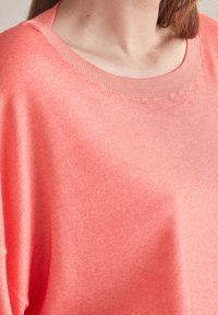 Kobieta w koralowo-różowym swetrze z okrągłym dekoltem, pokazana od szyi do połowy torsu, z prostymi brązowymi włosami widocznymi z prawej strony.