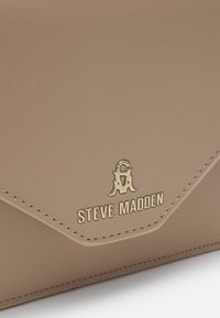 Sac à main Steve Madden couleur camel avec logo doré et nom de la marque sur un coin de rabat lisse et cousu. Aucune personne ni autre objet visible.