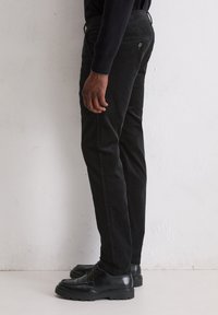 Pantalons en velours noir avec une coupe slim, poches latérales et une texture subtile. Associés à des chaussures en cuir noir, présentés de profil.