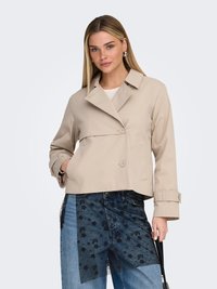 Vrouw draagt een beige cropped trenchcoat en patchwork jeans met zwarte bloemenprint, houdt een zwarte handtas vast tegen een witte achtergrond.