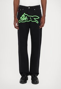 Black denim pants with a relaxed fit, featuring a large neon green graphic of a cartoon cat on the front. Black shoes complete the outfit.