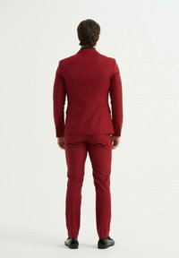 Costume rouge à coupe ajustée, veste à un bouton et pantalon slim. Revers en satin et poignets à trois boutons. Chaussures noires unies.