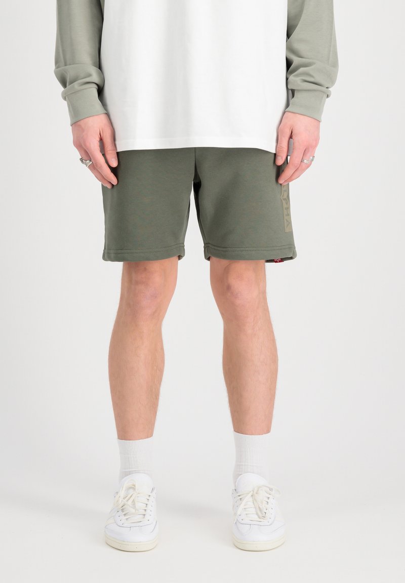 Shorts en coton vert olive avec une taille élastique, des poches latérales et un délicat logo. Associés à des baskets blanches et des chaussettes montantes.