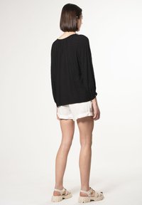 Schwarze Bluse mit langen Ärmeln und strukturiertem Muster; lockere Passform, runder Halsausschnitt, gerafftes Rückendesign. Getragen über weißen, gefalteten Shorts und beigen Sandalen.