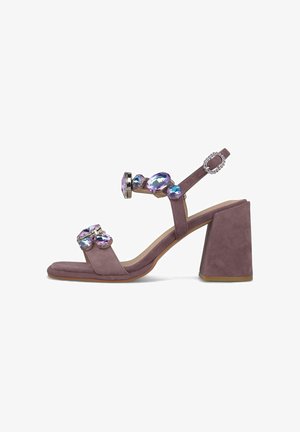 Sandalia de tacón alto de ante morado con tacón grueso tipo bloque, correa en el tobillo y grandes adornos de gemas iridiscentes en las correas y la hebilla.