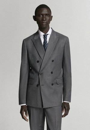 Hombre con traje gris cruzado con botones negros, camisa blanca y corbata azul marino a rayas, de pie frente a un fondo claro y liso.