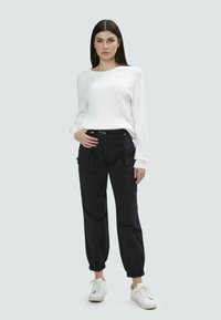 Weißer Pullover mit Kabelstrickmuster und Rundhalsausschnitt, kombiniert mit schwarzen Cargo-Hosen mit Taschen und elastischen Bündchen. Weiße Sneakers vervollständigen den Look.