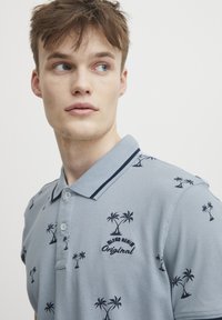 Lys blå polo t-skjorte med mørkeblå palmebroderi. Kragen har en marineblå stripe. Knappetrykke foran, korte ermer, teksturert stoff.