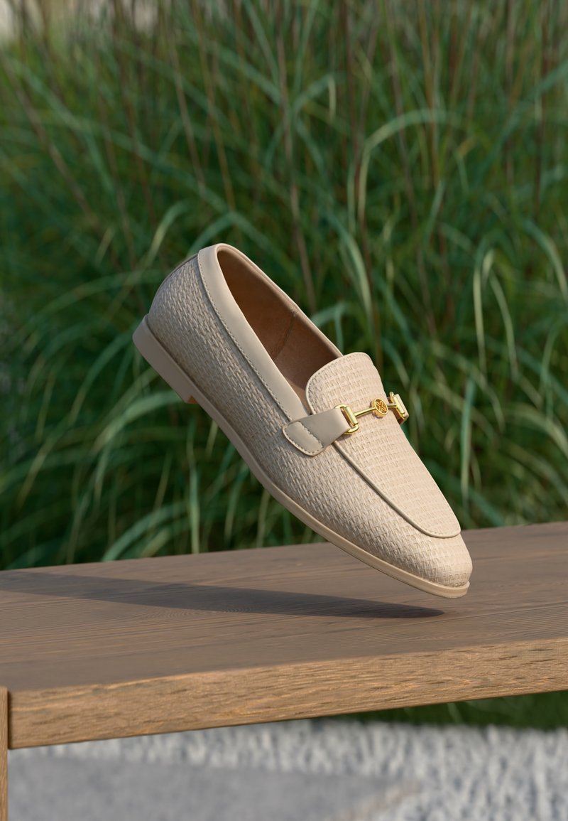 Mocassin en cuir tissé beige avec détail de boucle en métal doré, posé au-dessus d'un banc en bois avec un feuillage vert en arrière-plan.