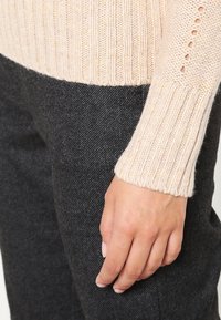 Beige gerippter Strickpulloverärmel mit offener Maschenstruktur am Handgelenk, der neben dunkelgrauen Wollhosen liegt.