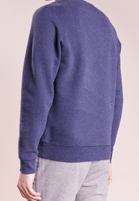 Hombre con un suéter de manga larga azul marino y pantalones gris claro, mostrado de espaldas contra un fondo claro.