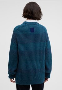 Υπερμεγέθη πλεκτή ζακέτα σε σκούρο τυρκουάζ με υφή ρίγας, που διαθέτει πιο σκούρες ρίγες και μια μικρή τετράγωνη ετικέτα στην πλάτη.